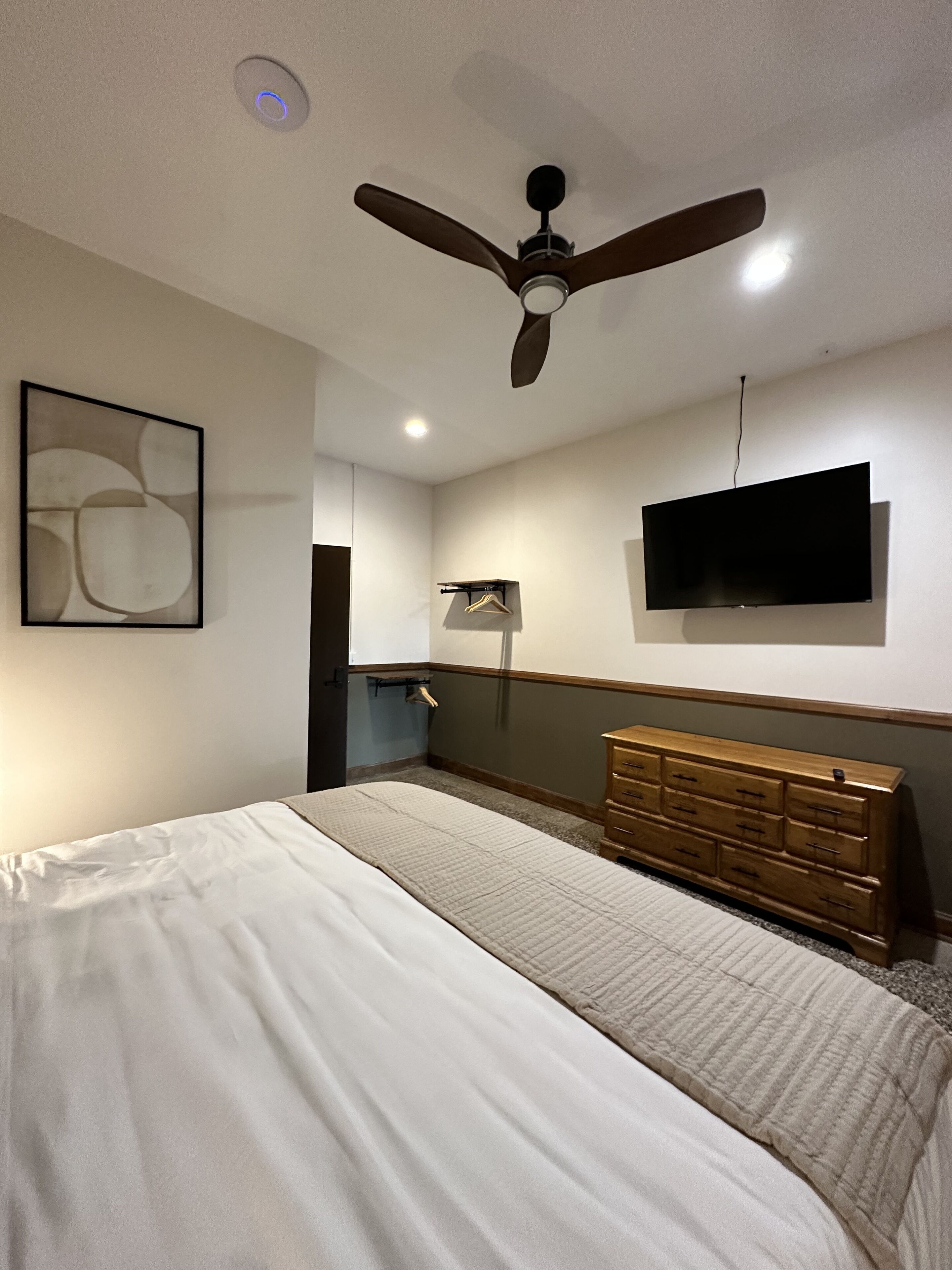 A bedroom with a bed, a dresser, a ceiling fan, a TV, and a wall-mounted shelf with clothes.