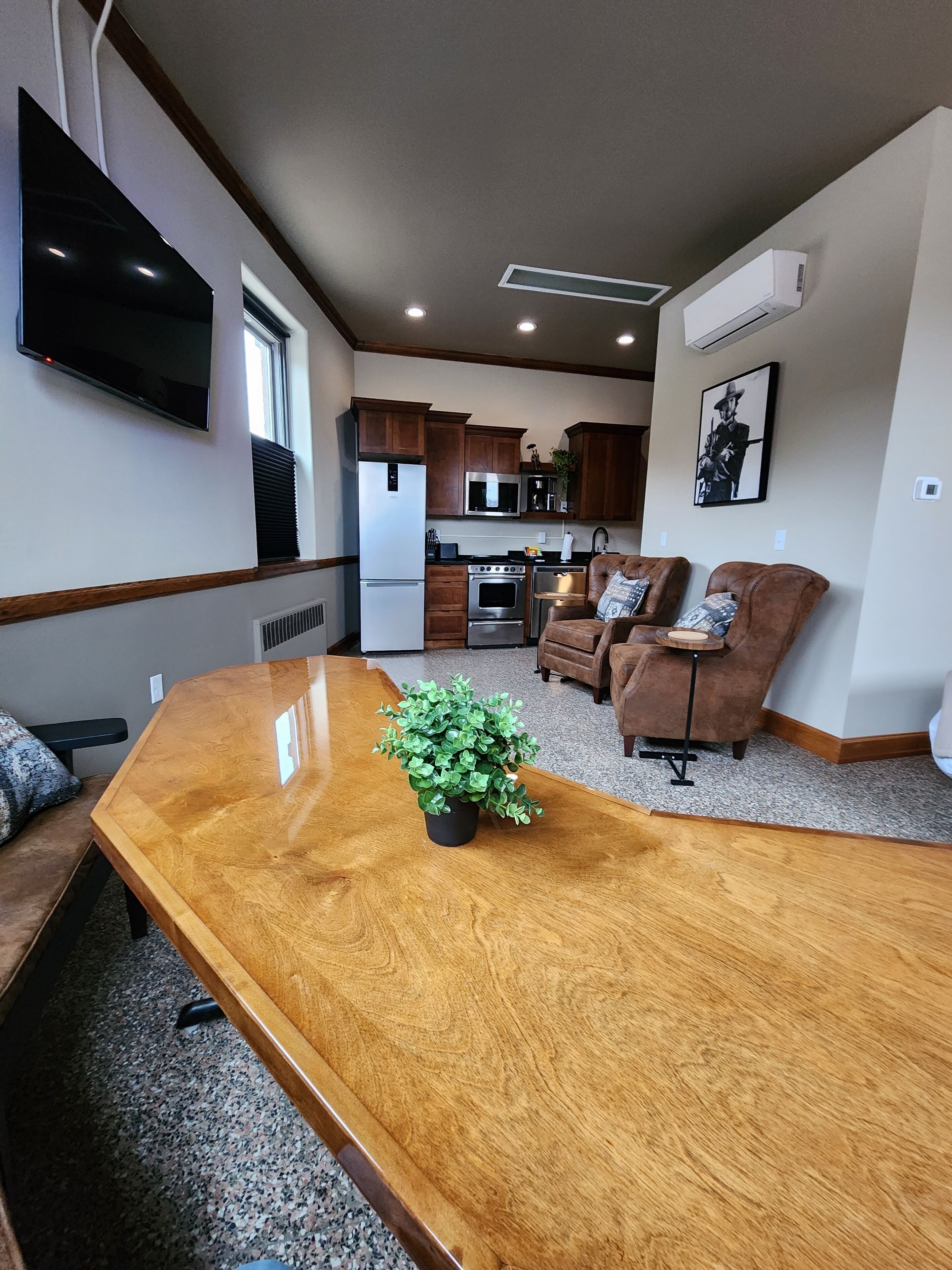 A modern living room with a wooden coffee table, two brown leather sofas, a flat-screen TV mounted on the wall, and a kitchen area in the background.