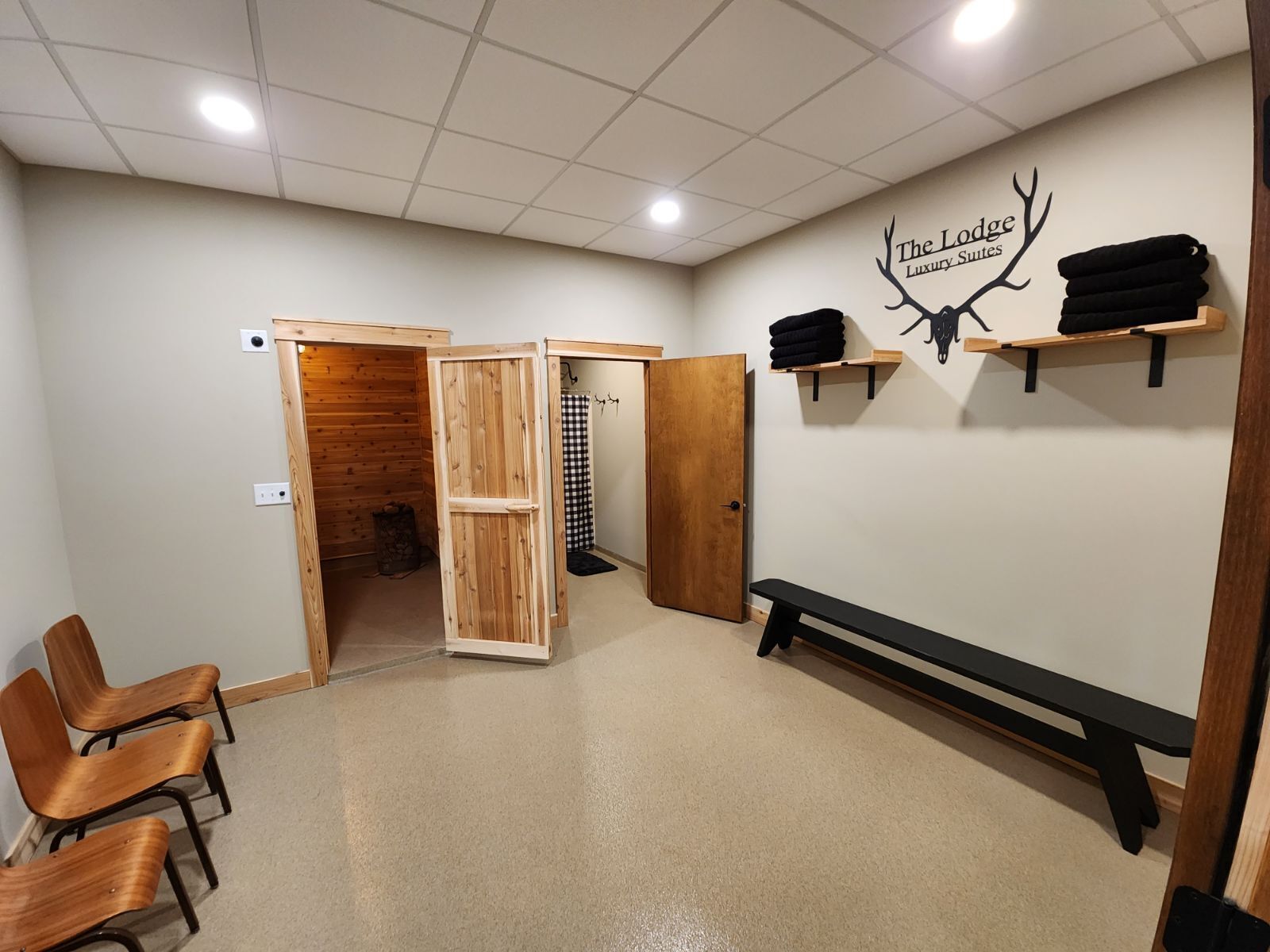 A room with a wooden bench, a bench with a black backrest, a wall-mounted shelf with towels, a door, and a wall decal of antlers with the text "The Lodge Luxury Suites".
