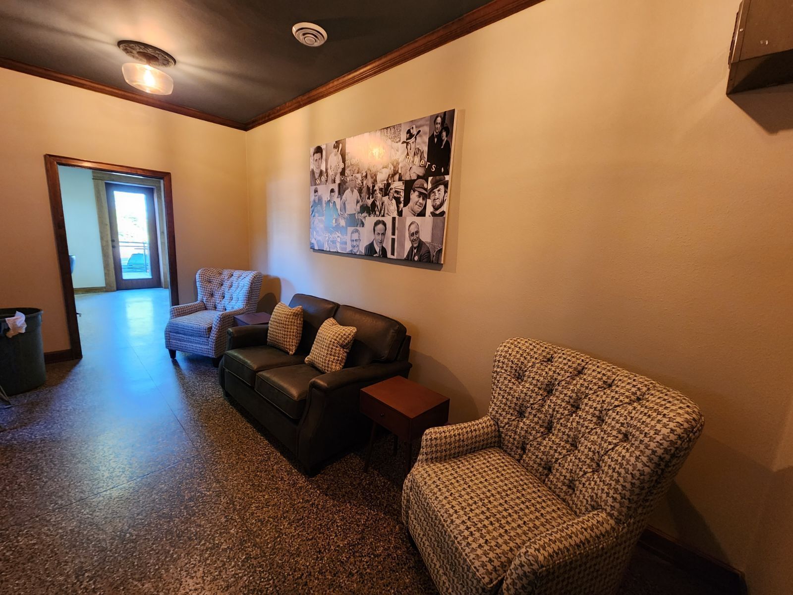 A living room with a black leather sofa, a patterned armchair, and a houndstooth armchair. A large collage of black and white photos adorns the wall.