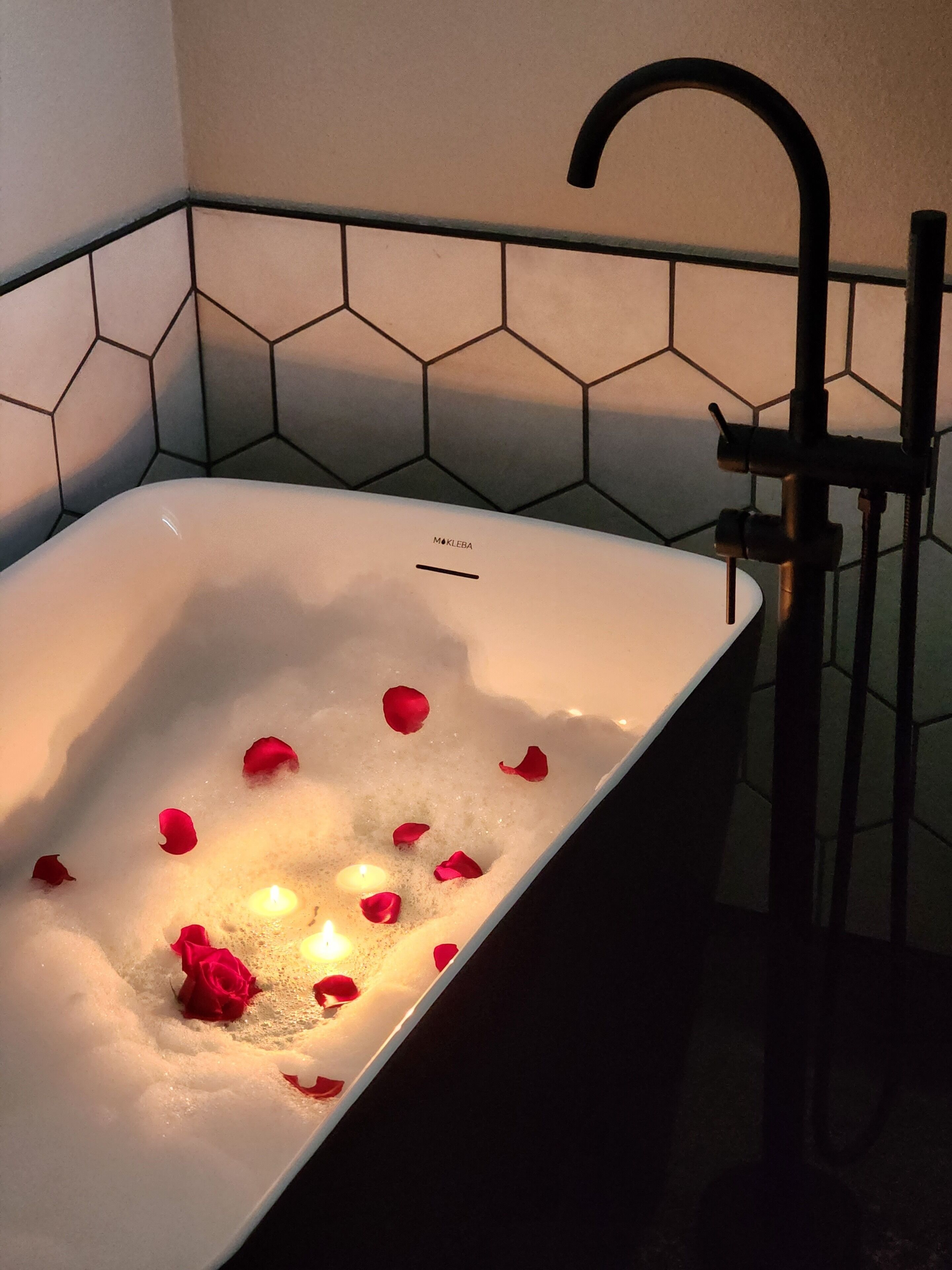 A bathtub with rose petals and a candle, a black faucet, and hexagonal tile walls.