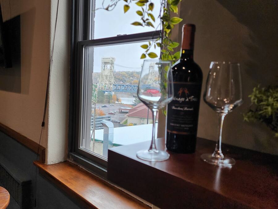 A window with a view of a bridge and buildings, a bottle of wine, and two wine glasses on a wooden ledge.