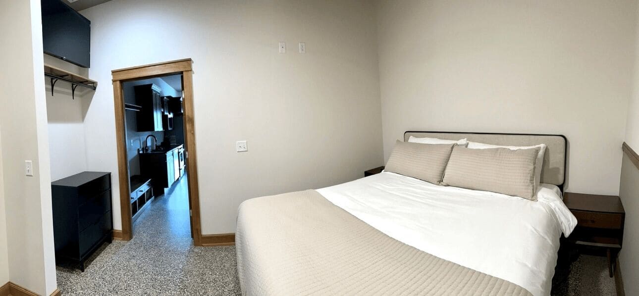 A bedroom with a bed, a nightstand, a wooden door leading to a kitchenette, and a small black cabinet.