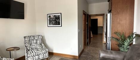 A living room with a flat-screen TV, patterned armchairs, a wooden coffee table with a plant, a gray sofa, and a kitchen area in the background.
