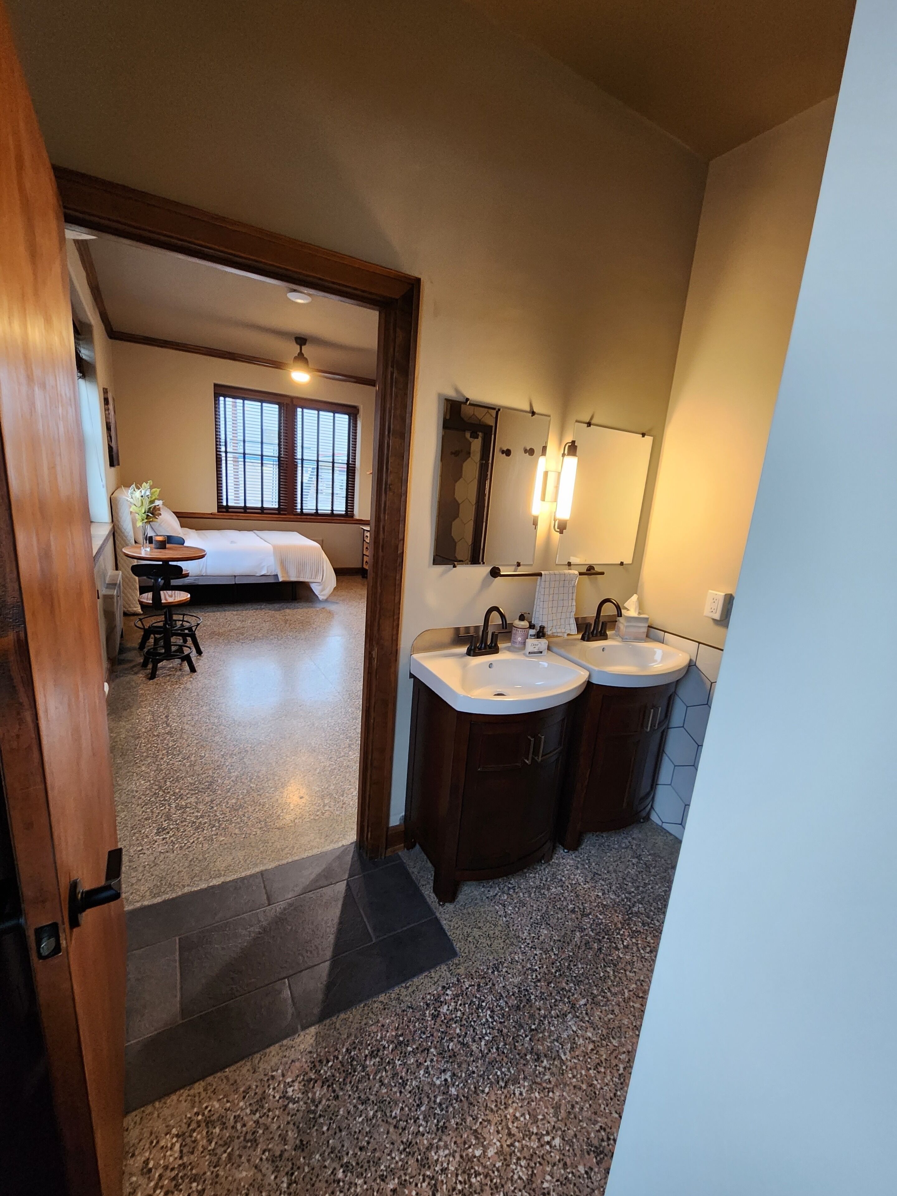 A hotel room with a bed, a small table with a vase, a bathroom with two sinks and a mirror, and a view of the outside through a window.