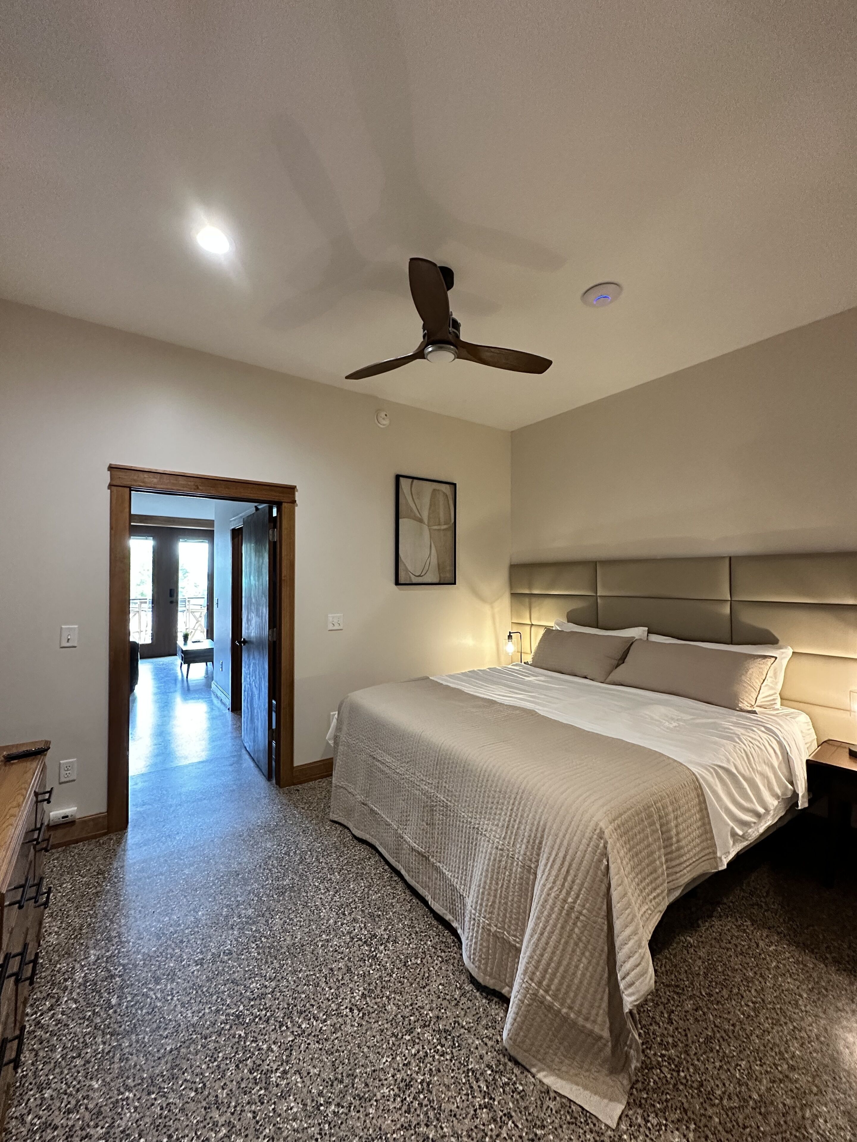 Une chambre à coucher comprenant un lit, un ventilateur de plafond, une table de chevet et un couloir menant à une autre pièce.