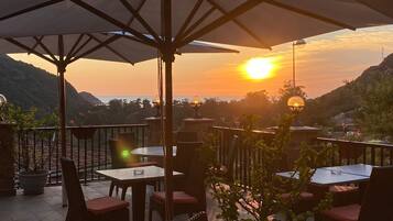 Une terrasse extérieure avec des tables et des chaises, des parasols et une vue sur le coucher de soleil se reflétant sur un paysage montagneux.