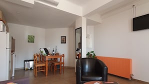 Un salon moderne comprenant un canapé, une table à manger avec des chaises, un fauteuil en cuir noir, une petite table avec un livre et une télécommande, et un téléviseur fixé au mur.