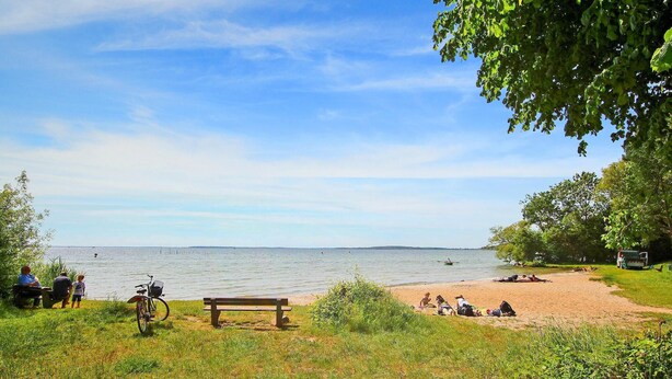 Un parc au bord du lac, avec un banc, un vélo et des gens qui se détendent sur le sable.
