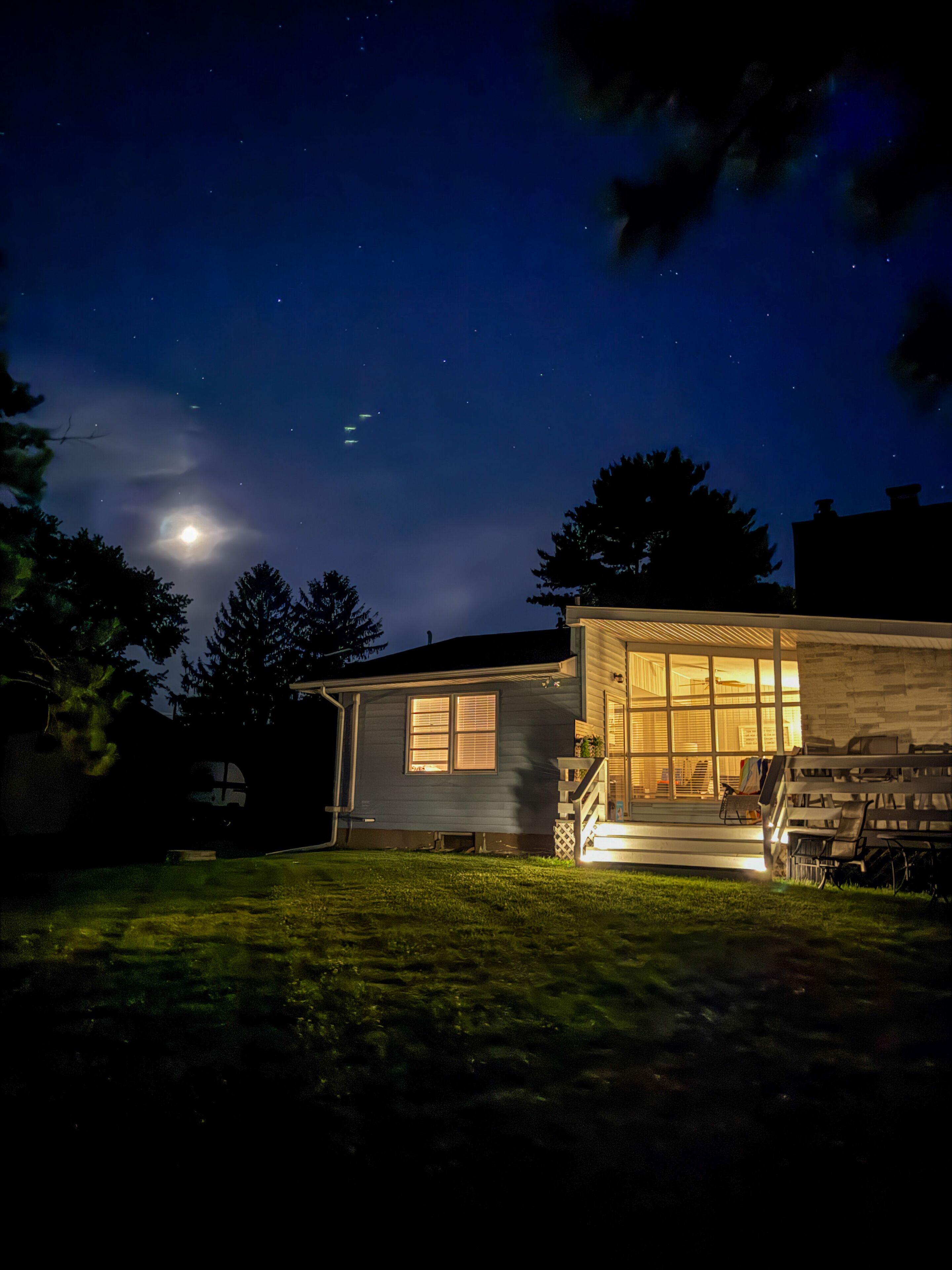 Ein Haus mit sichtbarer Veranda und einem gut beleuchteten Fenster, umgeben von einem dunklen Nachthimmel mit Sternen.