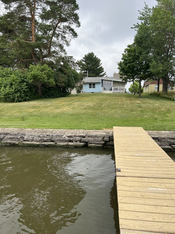 Una propiedad residencial con jardín césped, un muelle de madera que se adentra en un cuerpo de agua, y una casa al fondo.