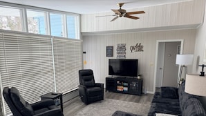 A living room with a black leather recliner, a black sofa, a TV on a black stand, a ceiling fan, and a window with blinds. - Cozy 3 bedroom, 1 bath home on Beaver Dam Lake (Beaver Dam)