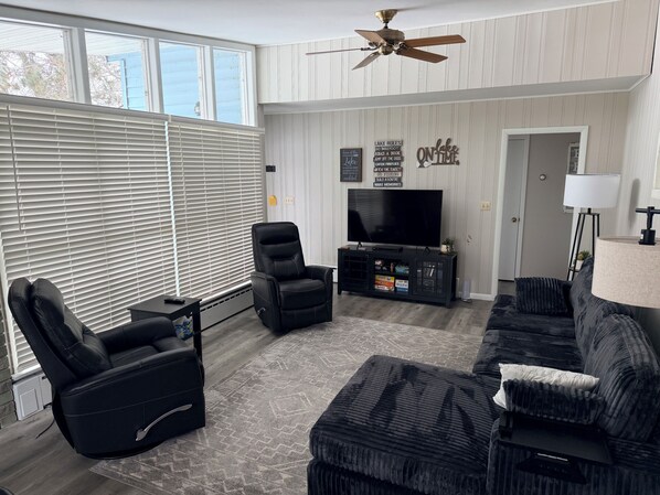 Un salon comprenant un fauteuil inclinable en cuir noir, un canapé noir, un téléviseur posé sur un meuble noir, un ventilateur de plafond et une fenêtre avec des stores.