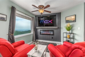 Una sala de estar con un sofá rojo, un ventilador de techo, un televisor de pantalla plana en la pared, una chimenea y una ventana con vistas a las montañas.