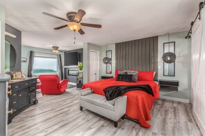 Un dormitorio con cama, una silla roja, un sofá gris, un ventilador de techo y una televisión.