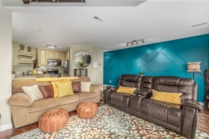 A living room with a brown leather sofa, a beige couch, and two ottomans. There is a kitchen in the background with a teal accent wall.