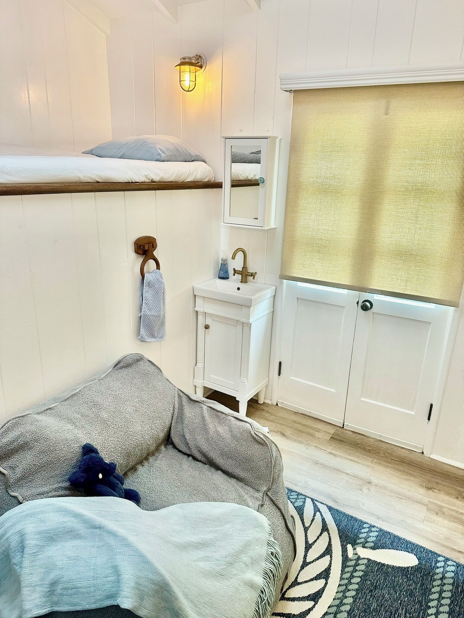 A room with a bed, a small white vanity with a mirror, a window with a curtain, and a blue patterned rug.