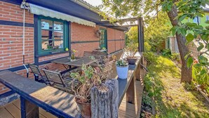 Ett hus byggt av tegel med en träterrass, en pergola och olika sorters krukväxter.