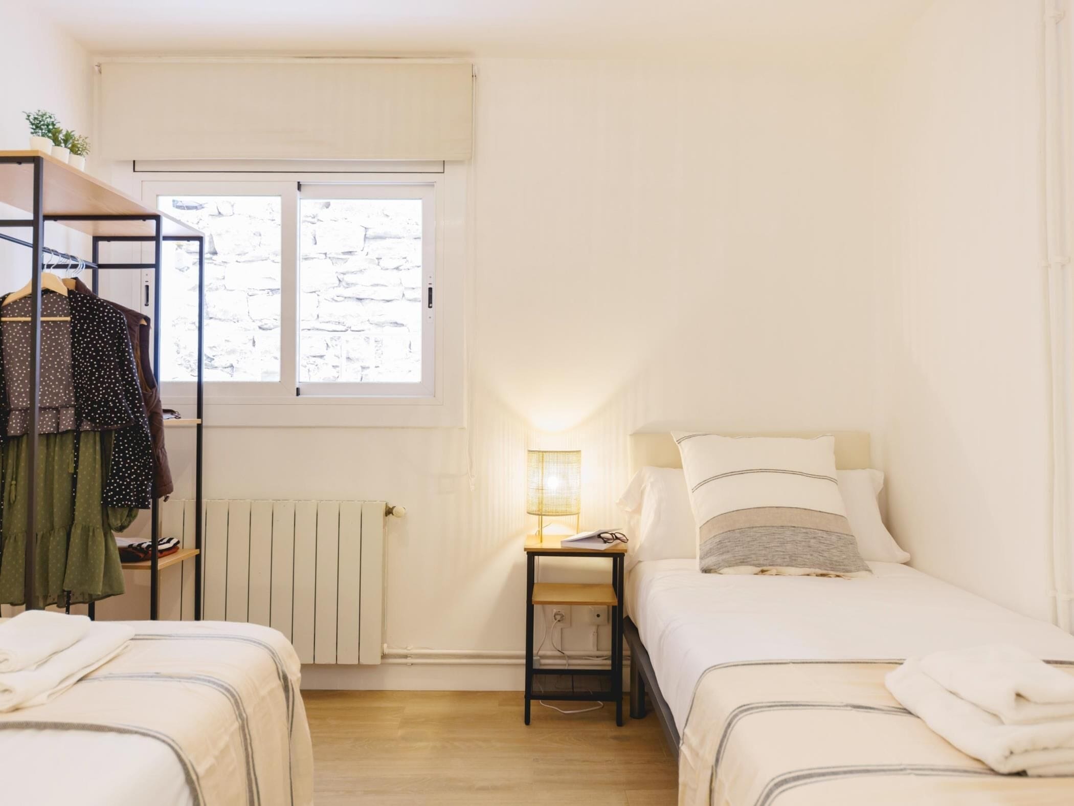 Et lille, godt oplyst soveværelse med seng, et sengebord med lampe, en radiator og et vindue med udsigt til en stenvæg.
