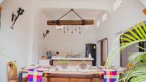 Un comedor con una mesa de madera, sillas y un mantel colorido. Hay plantas en macetas sobre la mesa y una lámpara colgante encima.