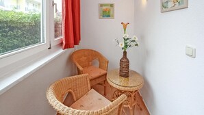 Una habitación de esquina con dos sillones de mimbre, una mesita redonda, un florero con flores, una ventana con cortinas rojas y una lámpara fijada a la pared.