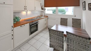 Una cocina con armarios blancos, encimera de madera, horno empotrado y comedor con sillas de mimbre.