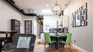 A modern kitchen with white cabinets, a dining area with a wooden table and green chairs, and a chandelier. - River Merchant Inn Mitchell’s Mercantile Suite (Stratford)