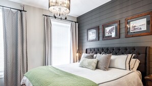 A bedroom with a large bed, grey curtains, a chandelier, and framed pictures on the wall. - River Merchant Inn Mitchell’s Mercantile Suite (Stratford)