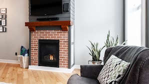 A living room with a brick fireplace, a flat-screen TV mounted above it, a grey sofa with a patterned cushion, and a potted plant. - River Merchant Inn Mitchell’s Mercantile Suite (Stratford)