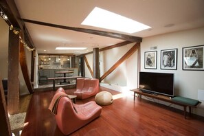 Un amplio salón con techo alto, vigas de madera, un ventanal grande, un televisor de pantalla plana y una zona de comedor al fondo.