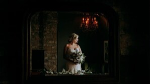 Una novia con vestido blanco sostiene un ramo, de pie en una habitación poco iluminada con pared de ladrillo y un candelabro.