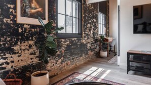 Une pièce avec des murs en briques apparentes, une grande fenêtre, une plante en pot, un bureau et un téléviseur.