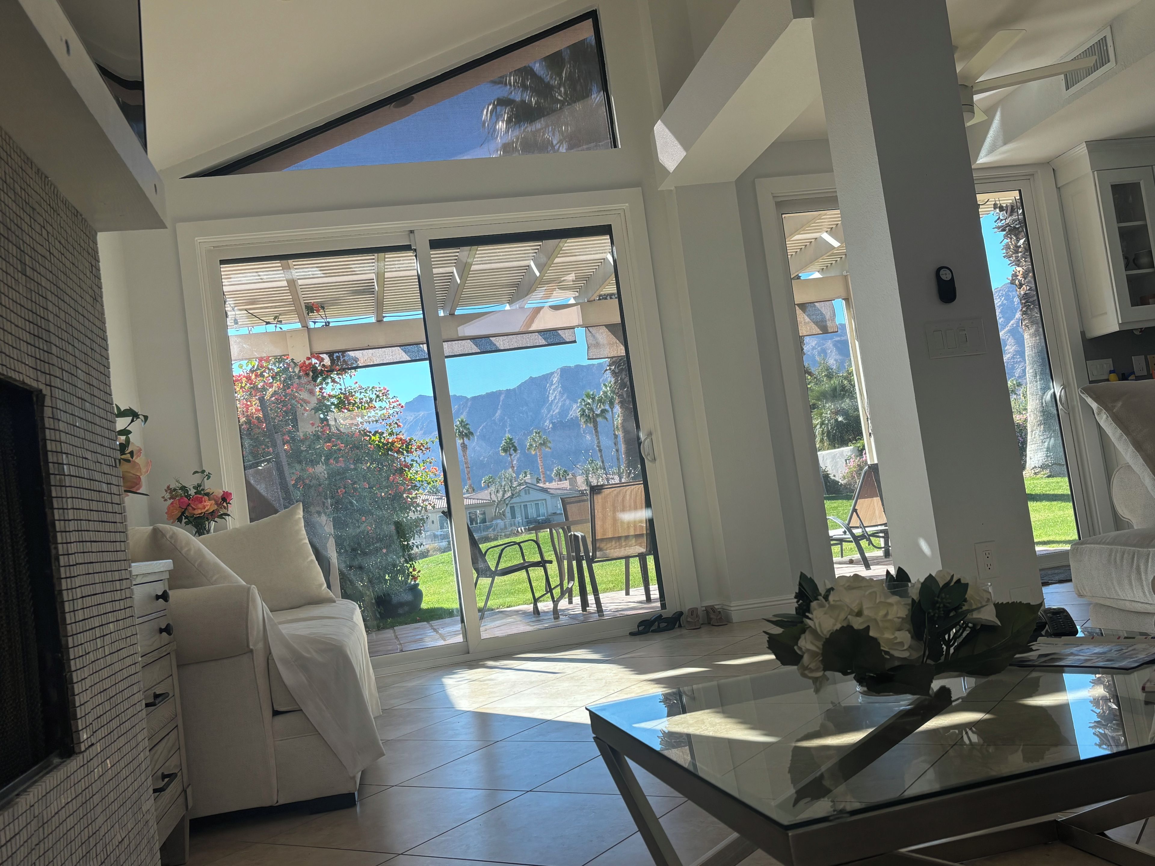 A modern living room with a glass table, white sofa, and a view of a garden and mountains.