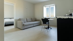 A modern, minimalistic living room with a sofa, a dining table with two chairs, a window with a view, and a door leading to another room.