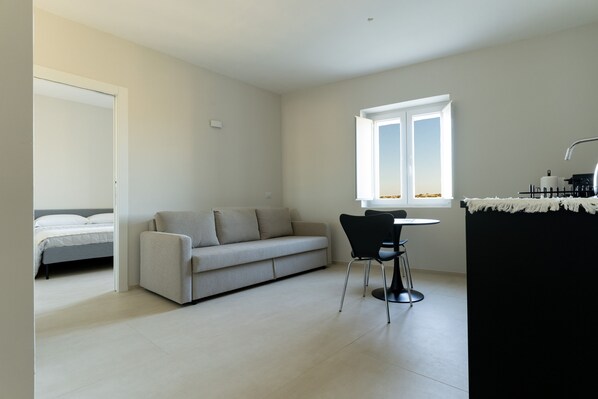 Un salon moderne et minimaliste, comprenant un canapé, une table à manger avec deux chaises, une fenêtre offrant une vue agréable et un lit dans la pièce adjacente.