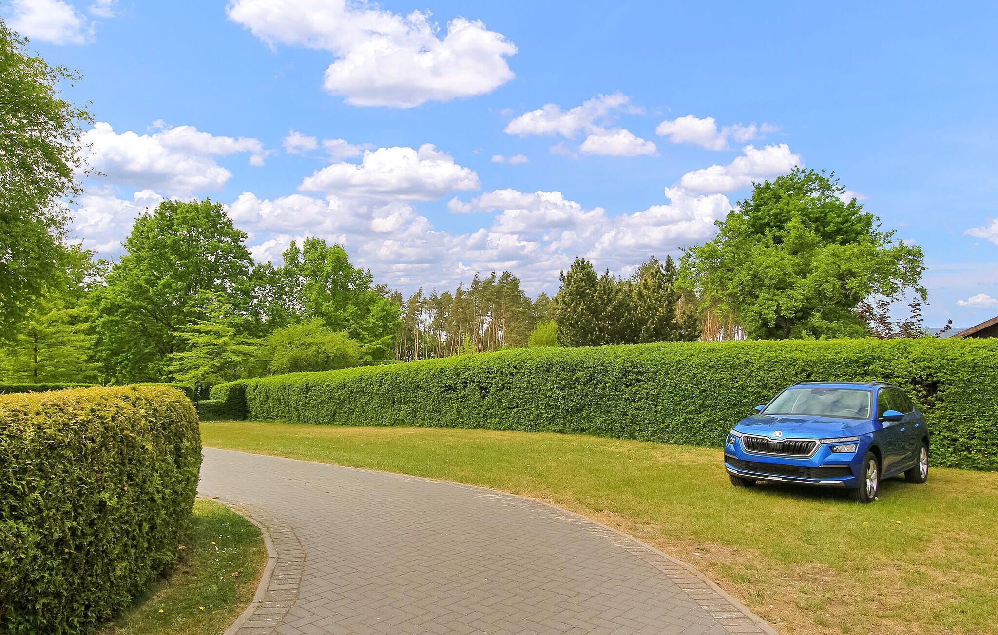 Un'auto blu parcheggiata su un vialetto lastricato in un'area curata con siepi e alberi sullo sfondo.