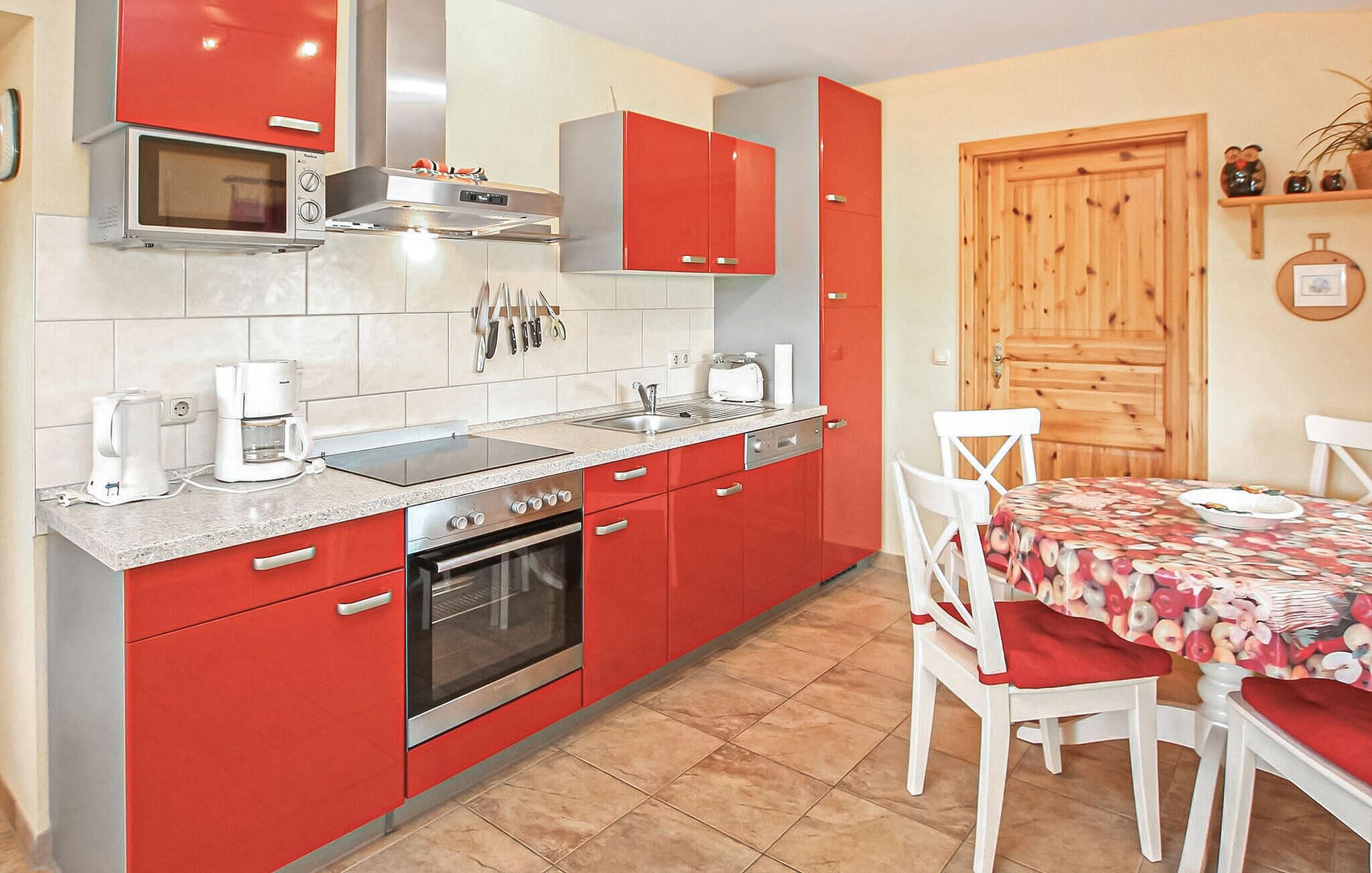 A kitchen with red cabinets, a built-in oven, a microwave, and a sink. There is a dining table with chairs and a wooden door in the background.