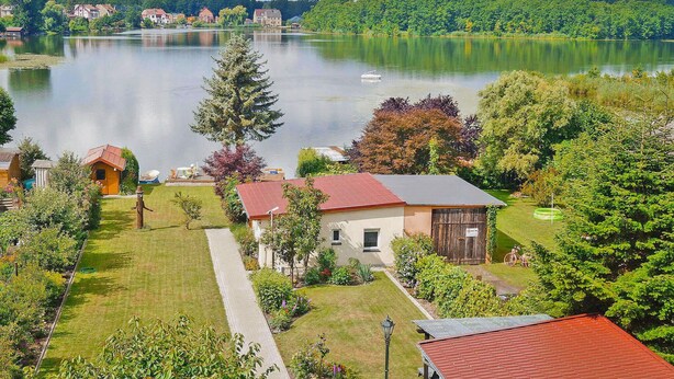 A lakeside property with a well-maintained lawn, a small building with a red roof, and a boat docked near the water. - Lovely home in Lychen with sauna (Lychen)