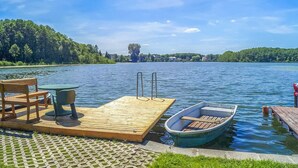 A wooden dock with a table and chairs, a small boat, and a clear view of a lake with trees and houses in the distance. - Lovely home in Lychen with sauna (Lychen)