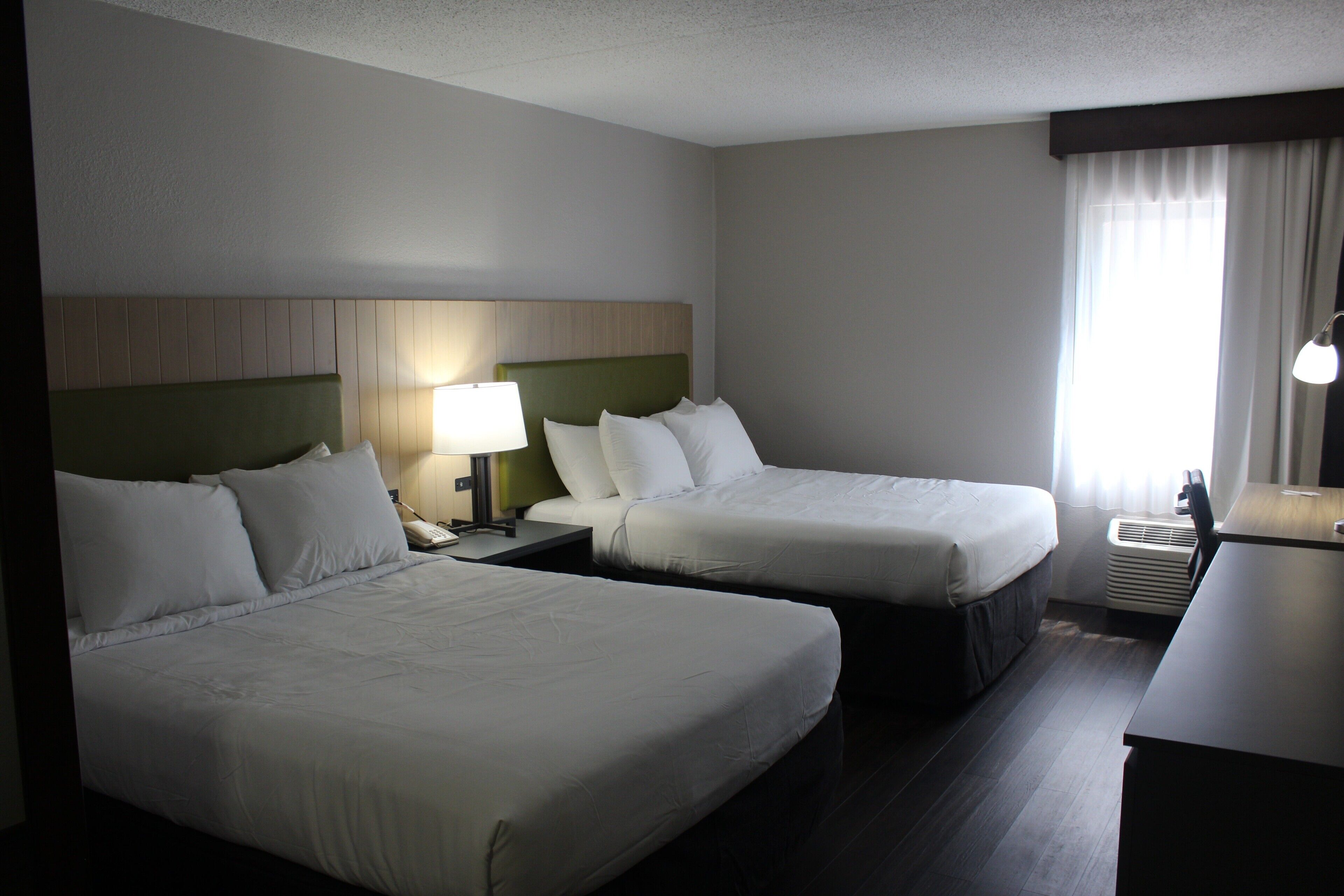 Habitación de hotel con dos camas, un escritorio, una lámpara y un ventanal con cortinas.
