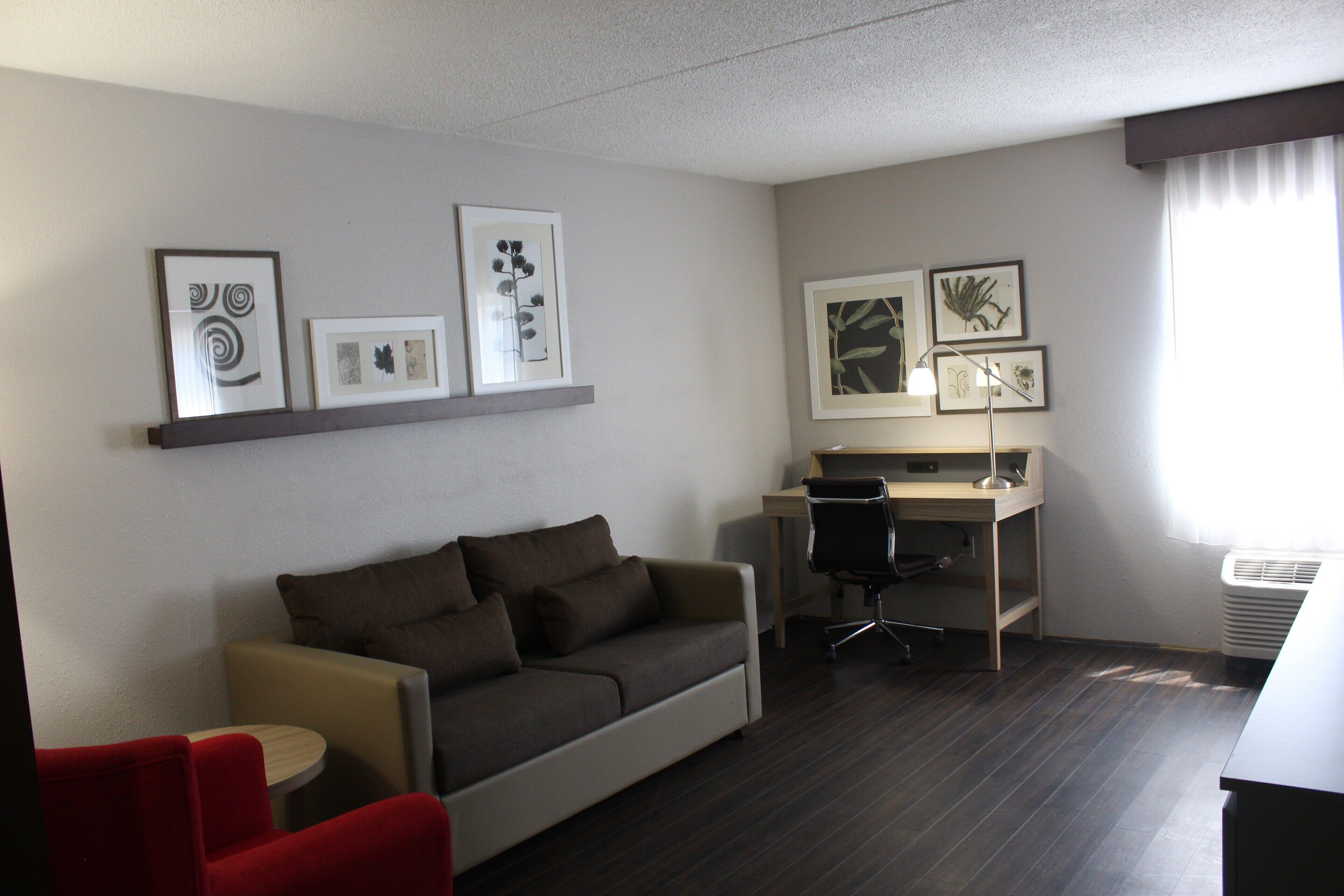 Une chambre d’hôtel moderne, équipée d’un canapé, d’un bureau et d’une chaise. Des tableaux encadrés ornent le mur, et une fenêtre est agrémentée de rideaux.