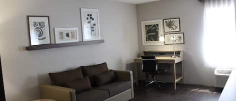 Ett modernt hotellrum med en soffa, ett skrivbord och en stol. På väggen hänger inramade tavlor och det finns ett fönster med gardiner.