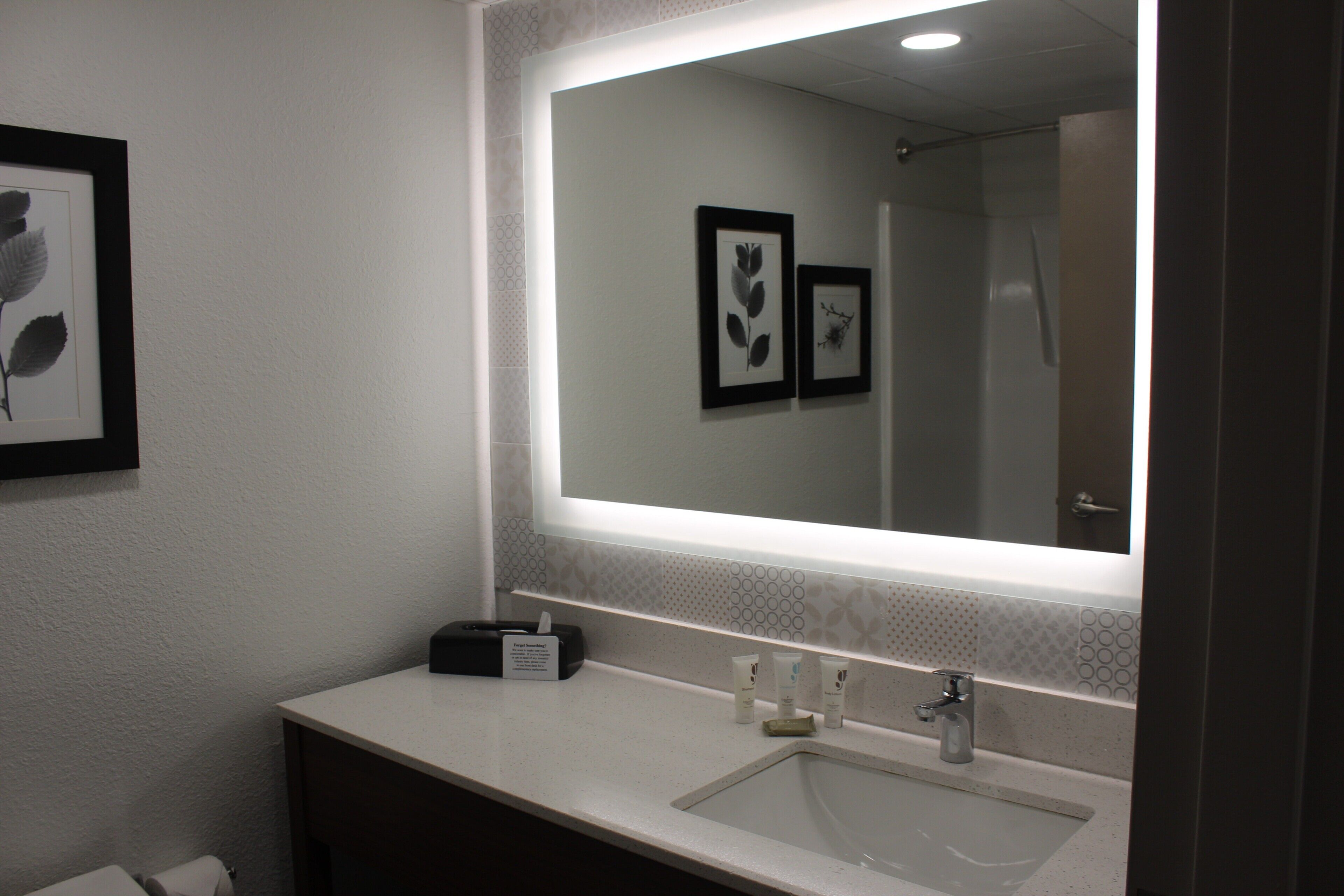 A bathroom with a large mirror, a sink, and a vanity. There are framed pictures on the wall and toiletries on the counter.