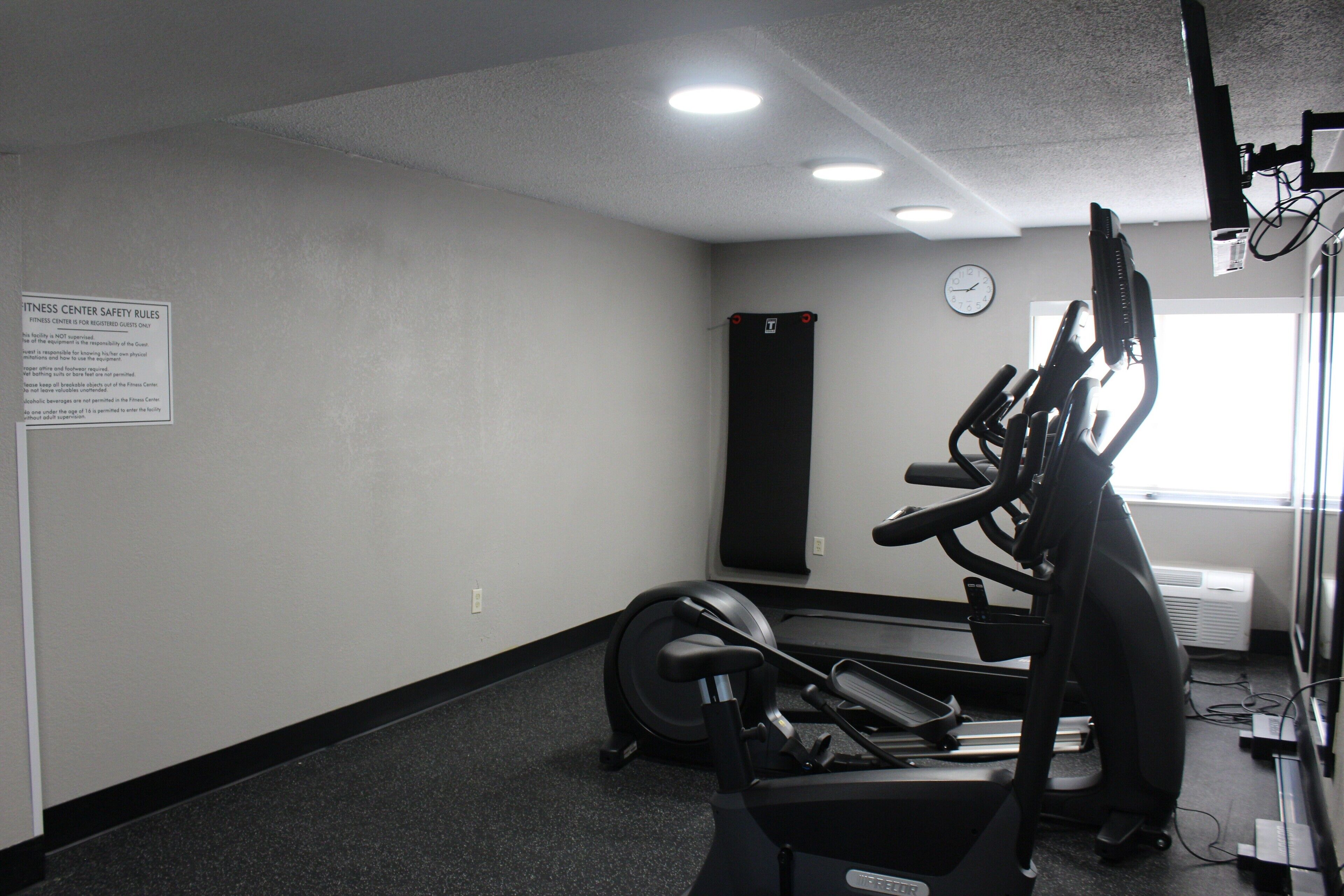 A fitness center with exercise equipment, a clock on the wall, and a sign with safety rules.