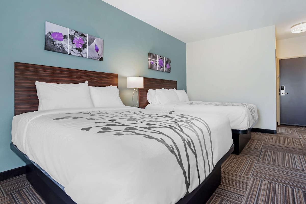 A hotel room with two beds, a headboard, a nightstand, a lamp, and wall art.