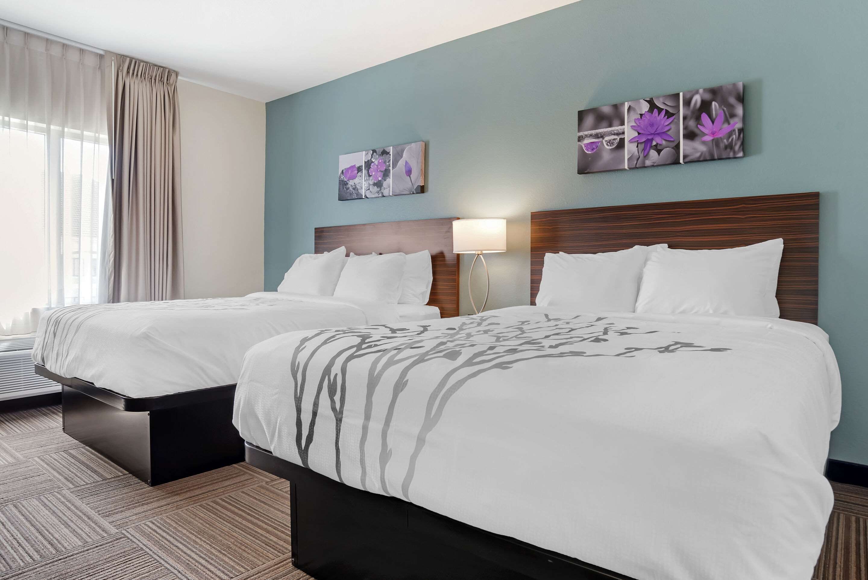 A hotel room with two beds, a wooden headboard, a wall-mounted lamp, and a window with curtains.