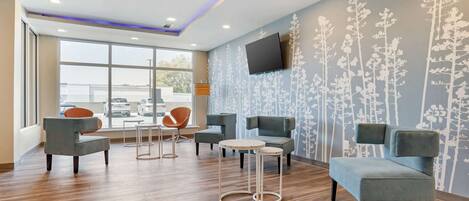 A modern lounge area with a wooden floor, grey upholstered seating, and a flat-screen TV mounted on the wall.