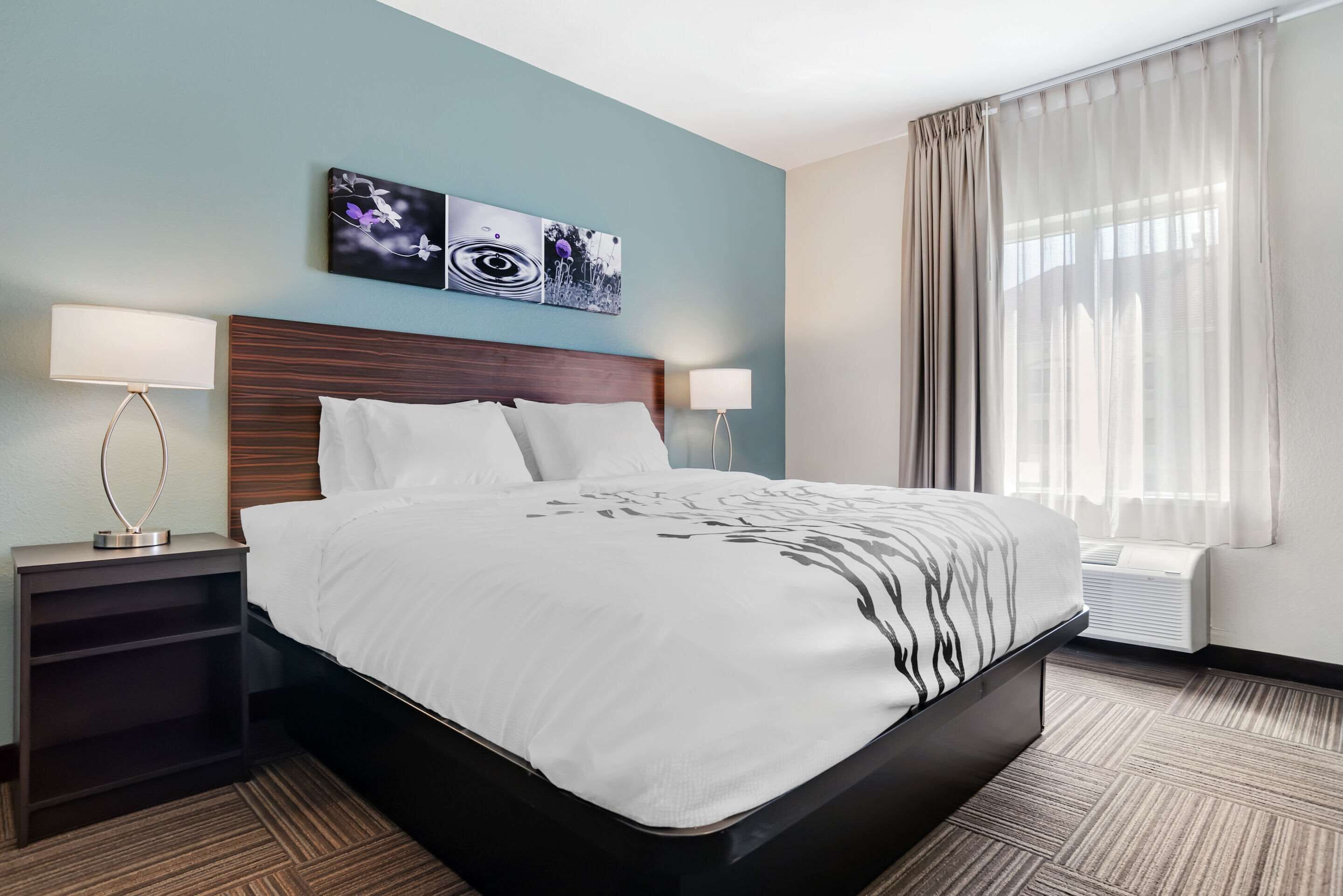 Quarto de hotel com uma cama grande, duas mesas de cabeceira com abajures, uma obra de arte na parede e uma janela com cortinas.
