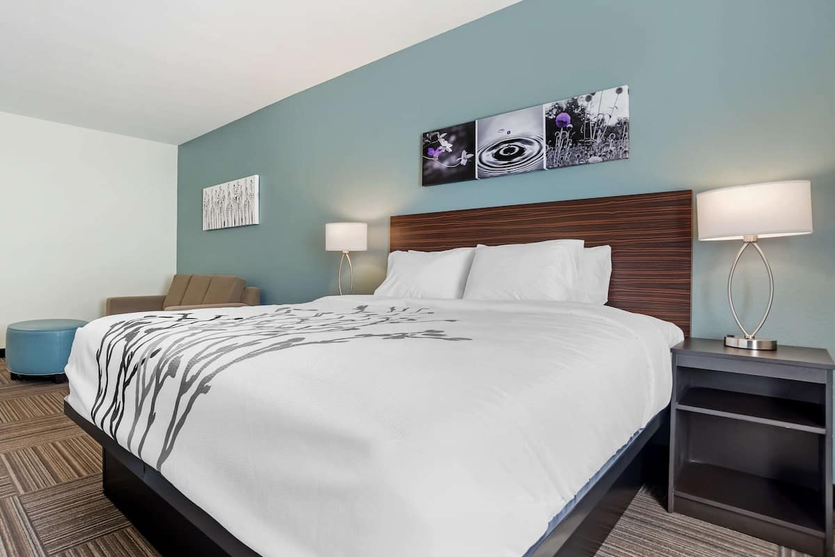 A modern hotel room with a large bed, two bedside tables with lamps, a blue ottoman, and artwork on the walls.