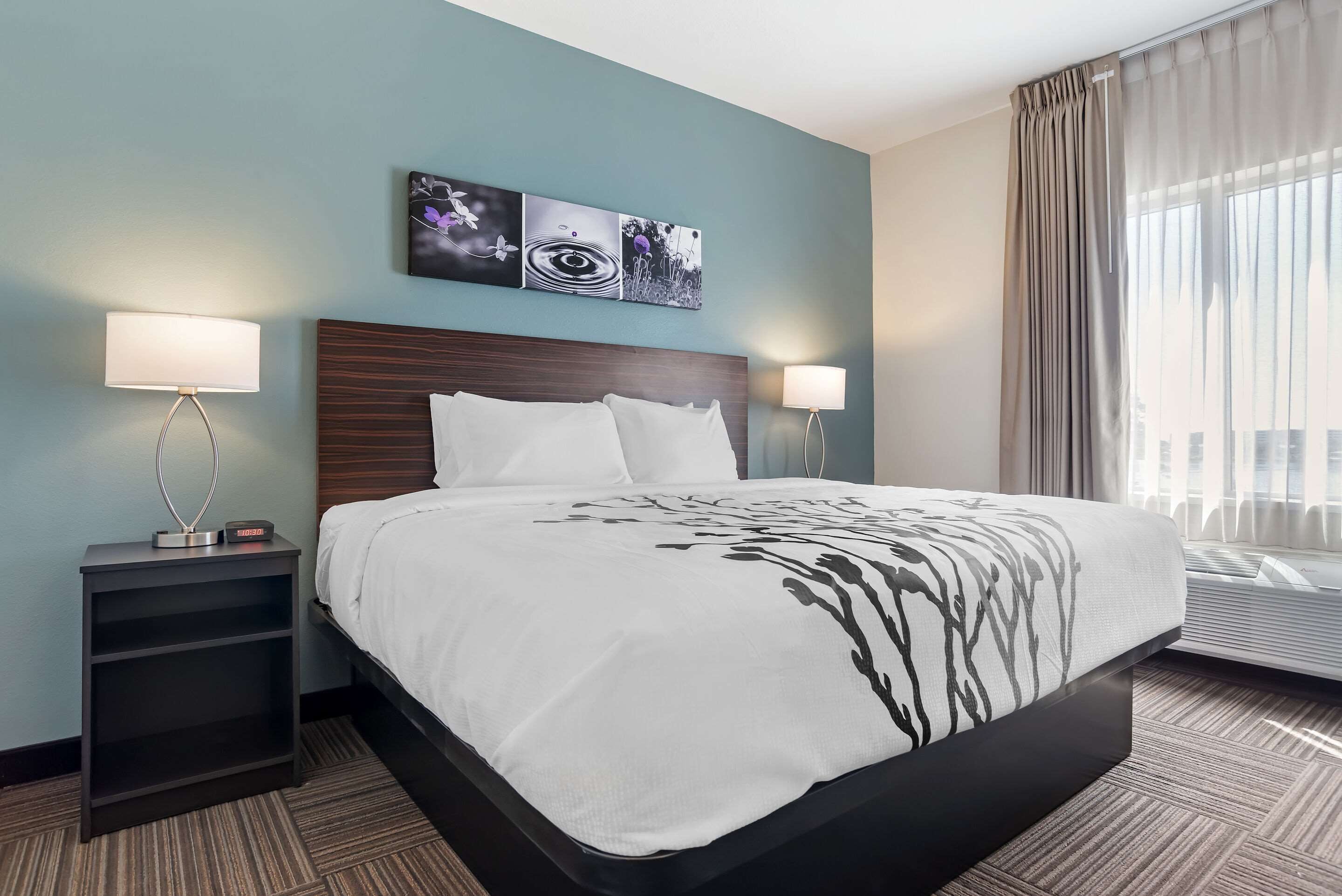 A hotel room with a bed, two bedside lamps, a nightstand, a wall art piece, and a window with curtains.